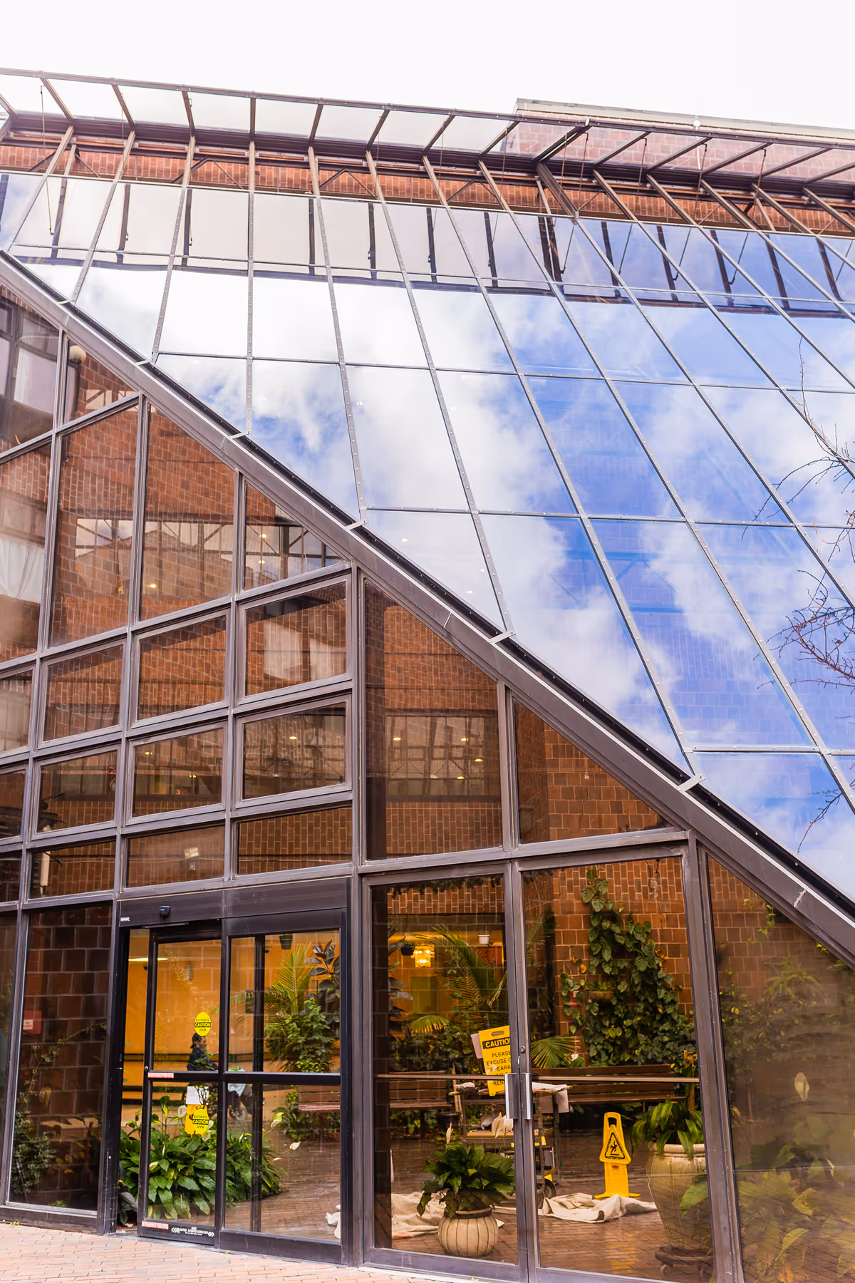 Exterior view of a building with large glass windows and a slanted glass roof reflecting the sky and clouds. The entrance has glass doors with plants visible inside and caution signs on the floor.