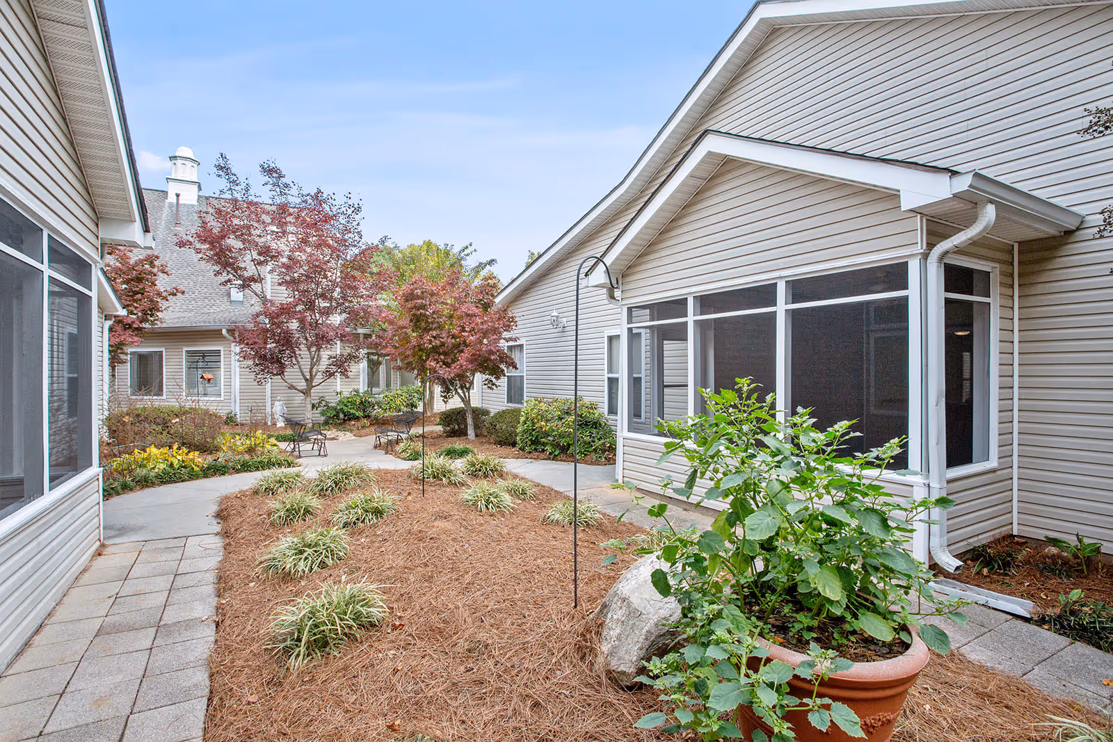 Outdoor courtyard area at Governors Glen featuring beige siding buildings, a paved walkway, landscaped garden beds with small bushes and plants, several red-leaved trees, and black metal benches.