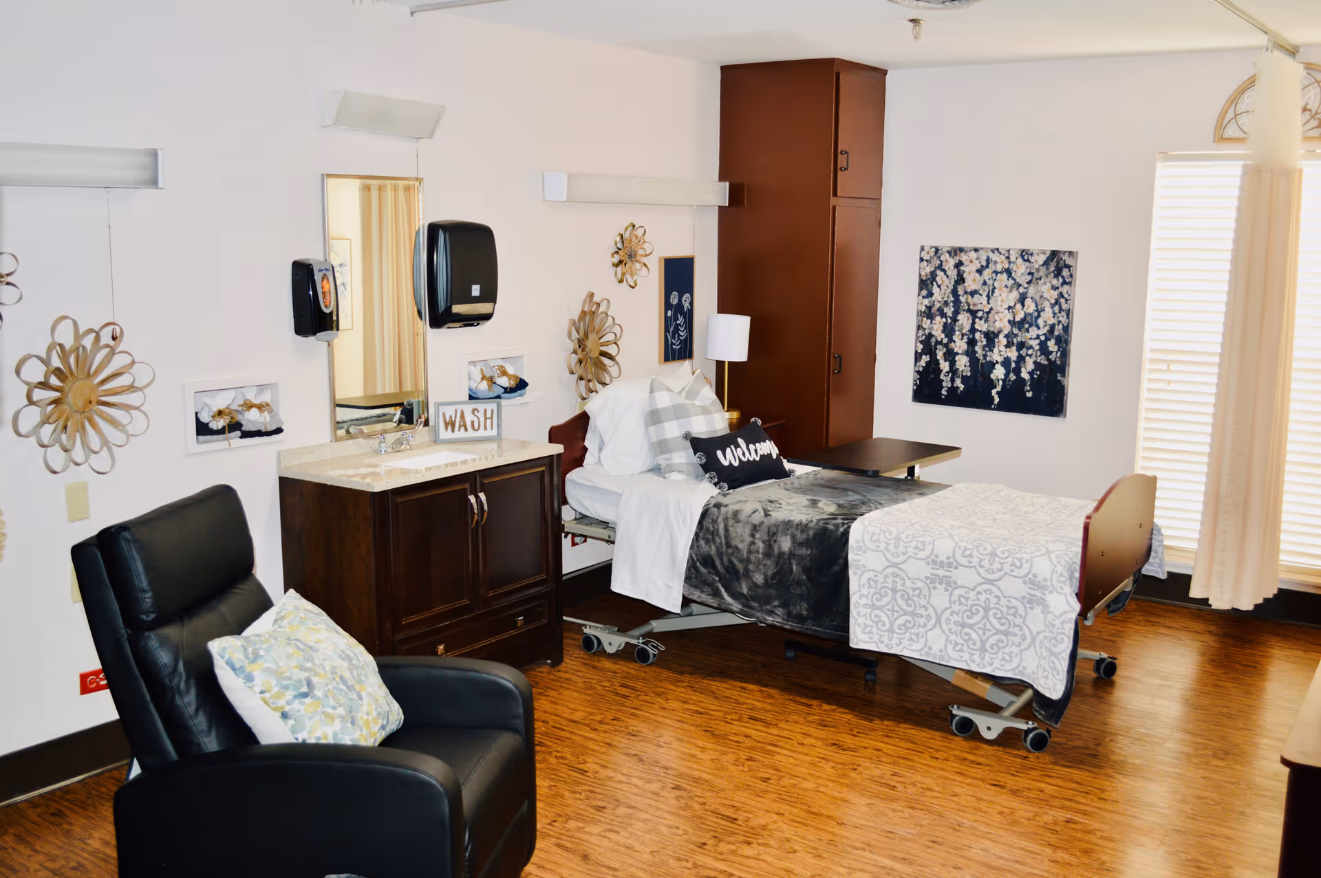 A well-lit senior living bedroom featuring a hospital-style bed with white and gray bedding, a black recliner chair with a patterned pillow, a wooden cabinet with a sink and a mirror above it, decorative wall art, and a window with blinds and curtains allowing natural light to enter.