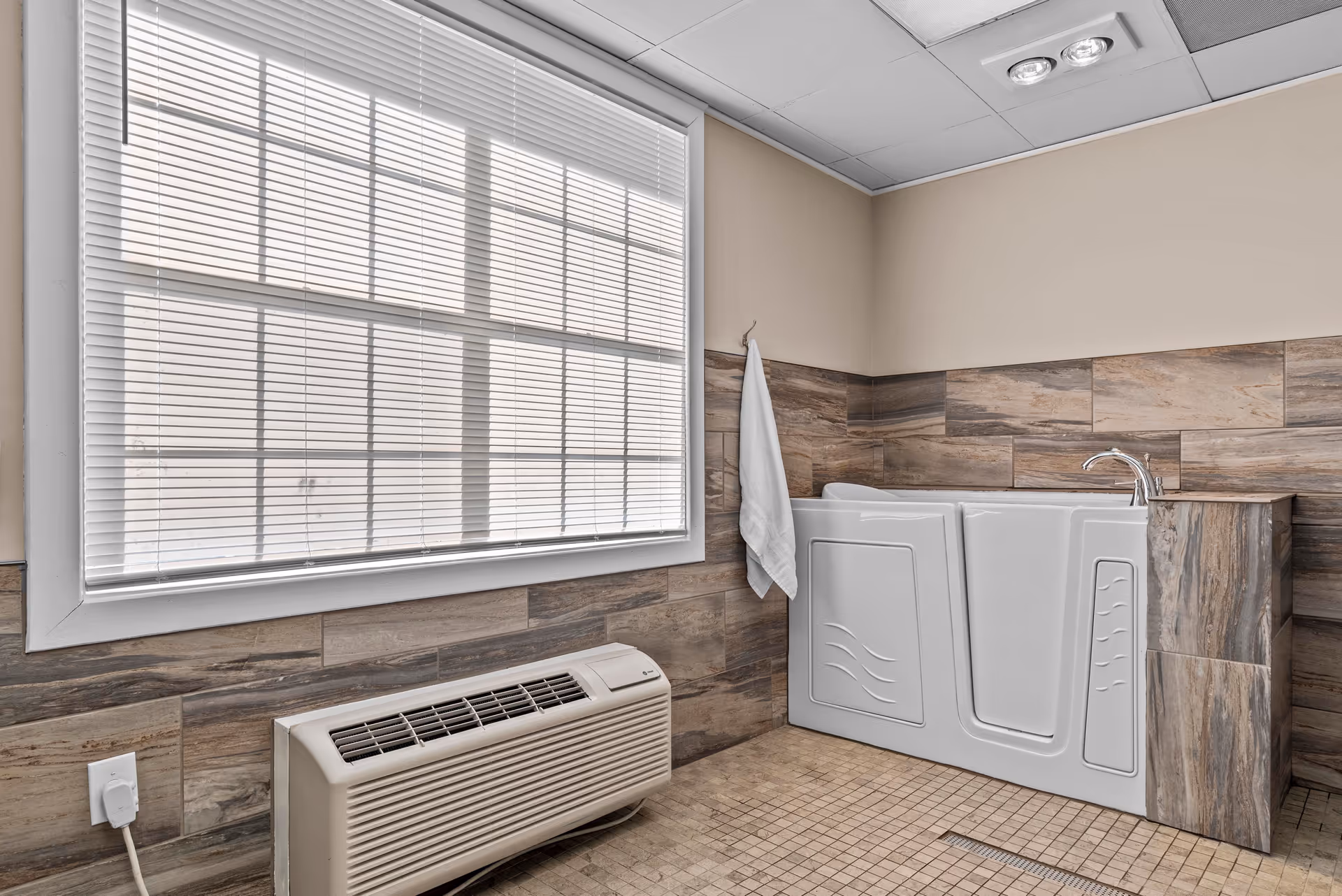 A walk-in bathtub with a door for easy access is installed in a bathroom with wood-patterned tile walls and floor. A white towel hangs on a hook next to the tub, and a large window with closed blinds is above a beige heating or air conditioning unit.