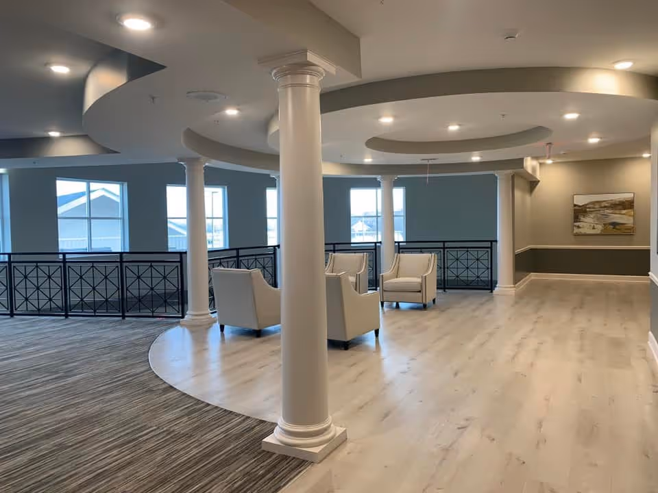 A spacious, modern interior common area with light wood flooring and carpeted sections. The space features white columns, several beige armchairs arranged in small groups, large windows letting in natural light, and a decorative ceiling with recessed lighting. A painting hangs on the far wall.