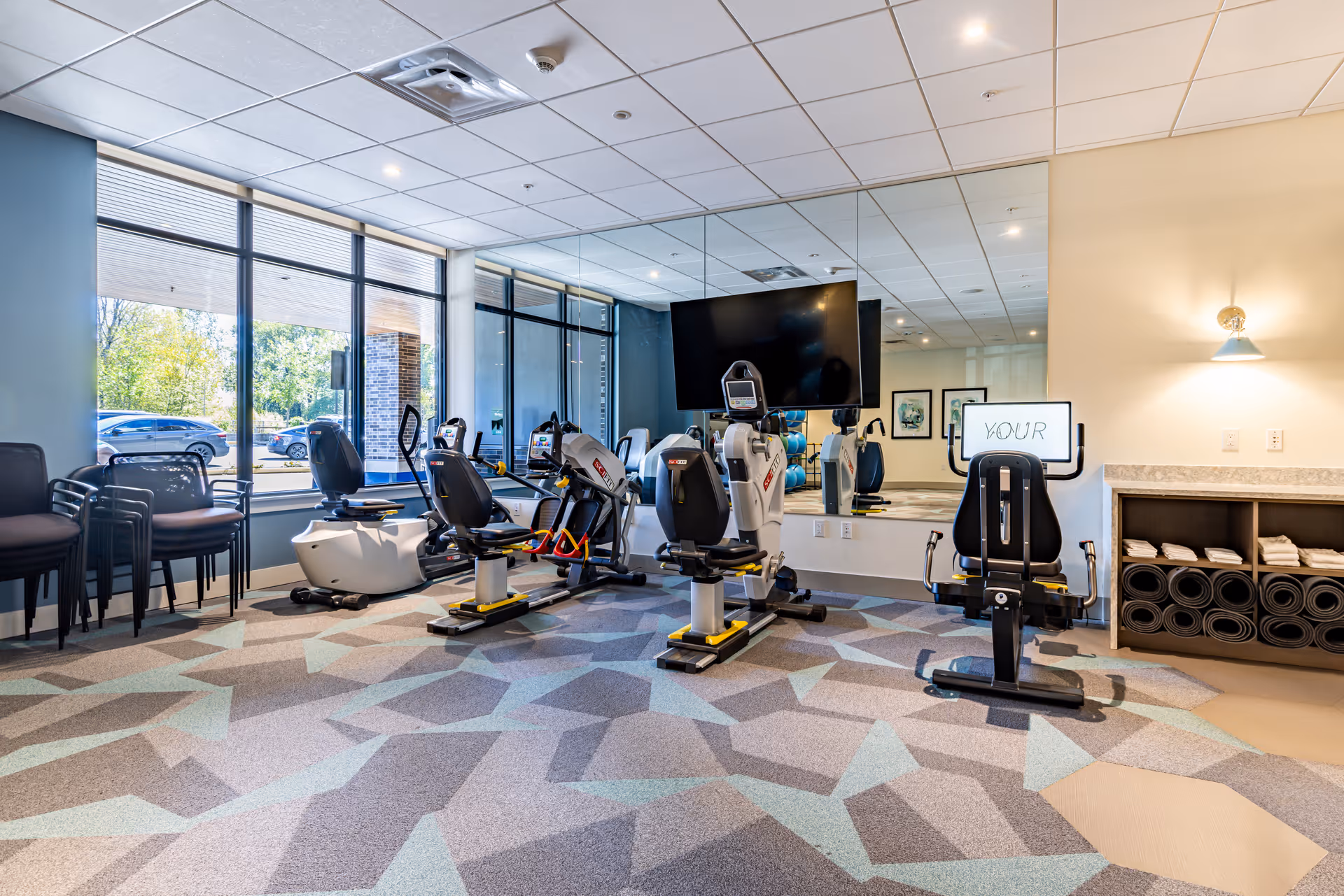 Well-lit indoor fitness room with exercise bikes and cardio machines, a mirrored wall, and stacked chairs by large windows.
