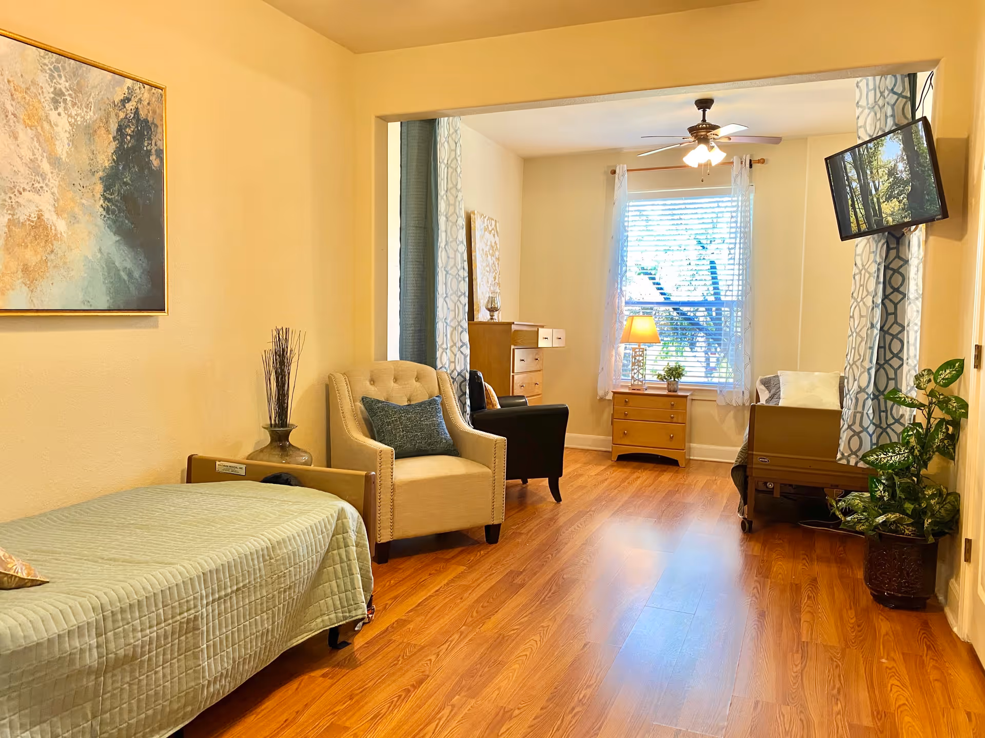 Bright assisted-living room with a daybed, upholstered chairs, wooden furniture, a wall-mounted TV, and a window letting in natural light.