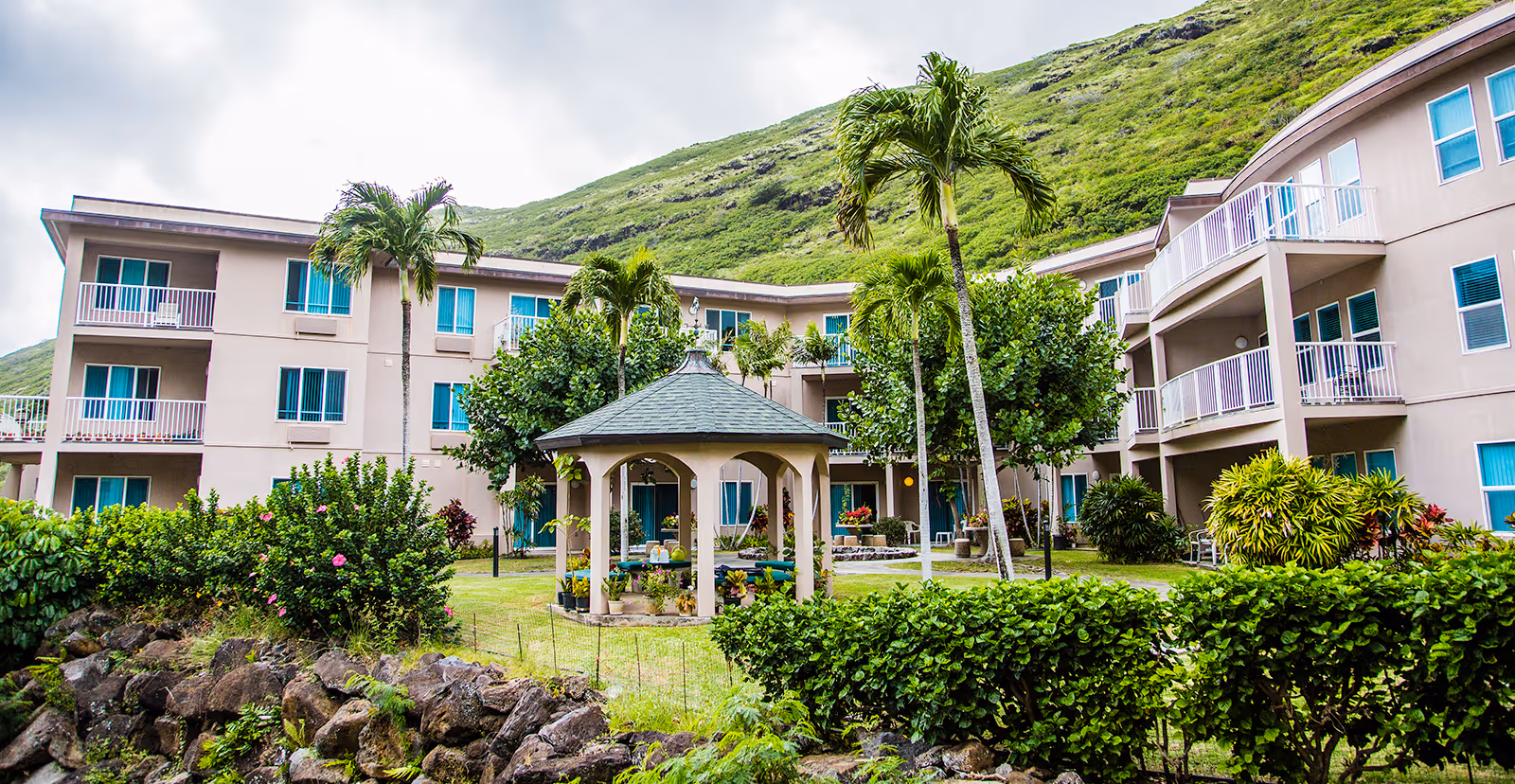 Outdoor courtyard area of The Ivy at Hawaii Kai senior living facility with a gazebo, palm trees, green bushes, and a three-story building in the background set against a green hillside under a cloudy sky.