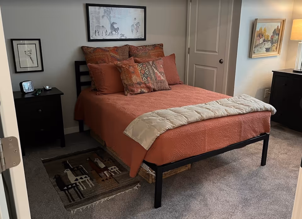 A neatly made bed with rust-colored bedding and patterned pillows in a furnished bedroom with nightstands and framed artwork on the walls.