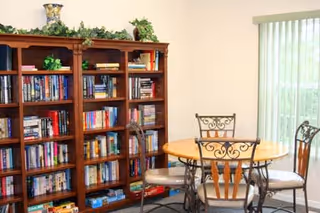 A cozy interior room featuring a wooden bookshelf filled with books and decorative plants on top, next to a round wooden table with four metal chairs with cushioned seats. A large window with vertical blinds allows natural light to enter the room.