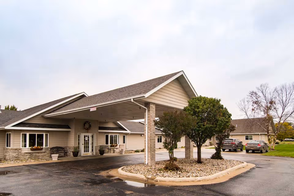 Exterior view of a single-story senior living facility building with a covered entrance supported by brick columns. There are a few trees and shrubs planted in a landscaped island in front of the entrance. Several cars are parked nearby, and the sky is overcast.