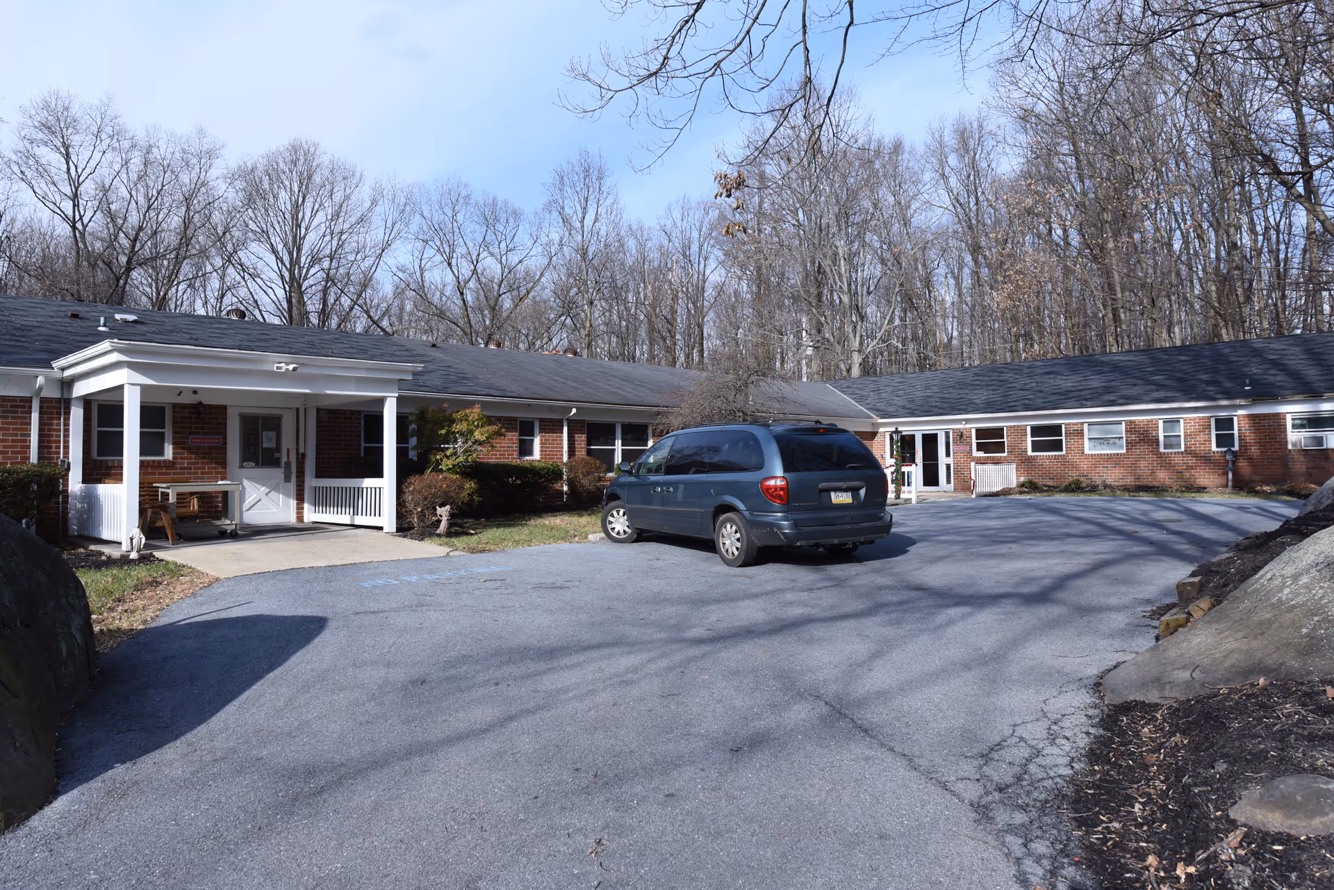 Low single-story brick nursing facility with a covered entrance, a parked minivan in the driveway, and leafless trees behind it.