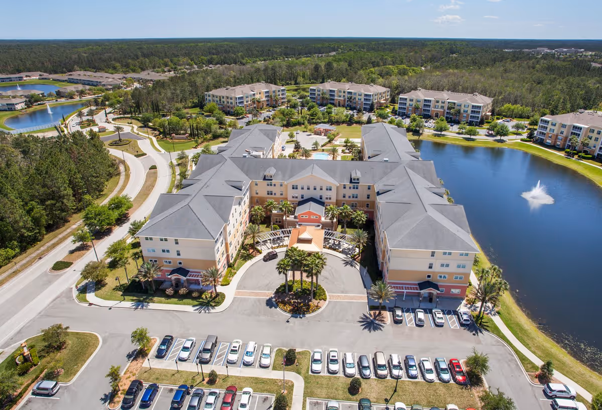 Aerial view of Camellia At Deerwood senior living facility showing a large multi-wing building surrounded by parking lots, landscaped gardens, a pond with a fountain, and nearby residential buildings under a clear blue sky.
