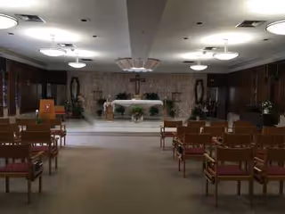 Interior view of a chapel or worship room with rows of wooden chairs with red cushions facing an altar decorated with flowers and a cross mounted on the back wall. The room has soft lighting from ceiling fixtures and plants on either side of the altar.