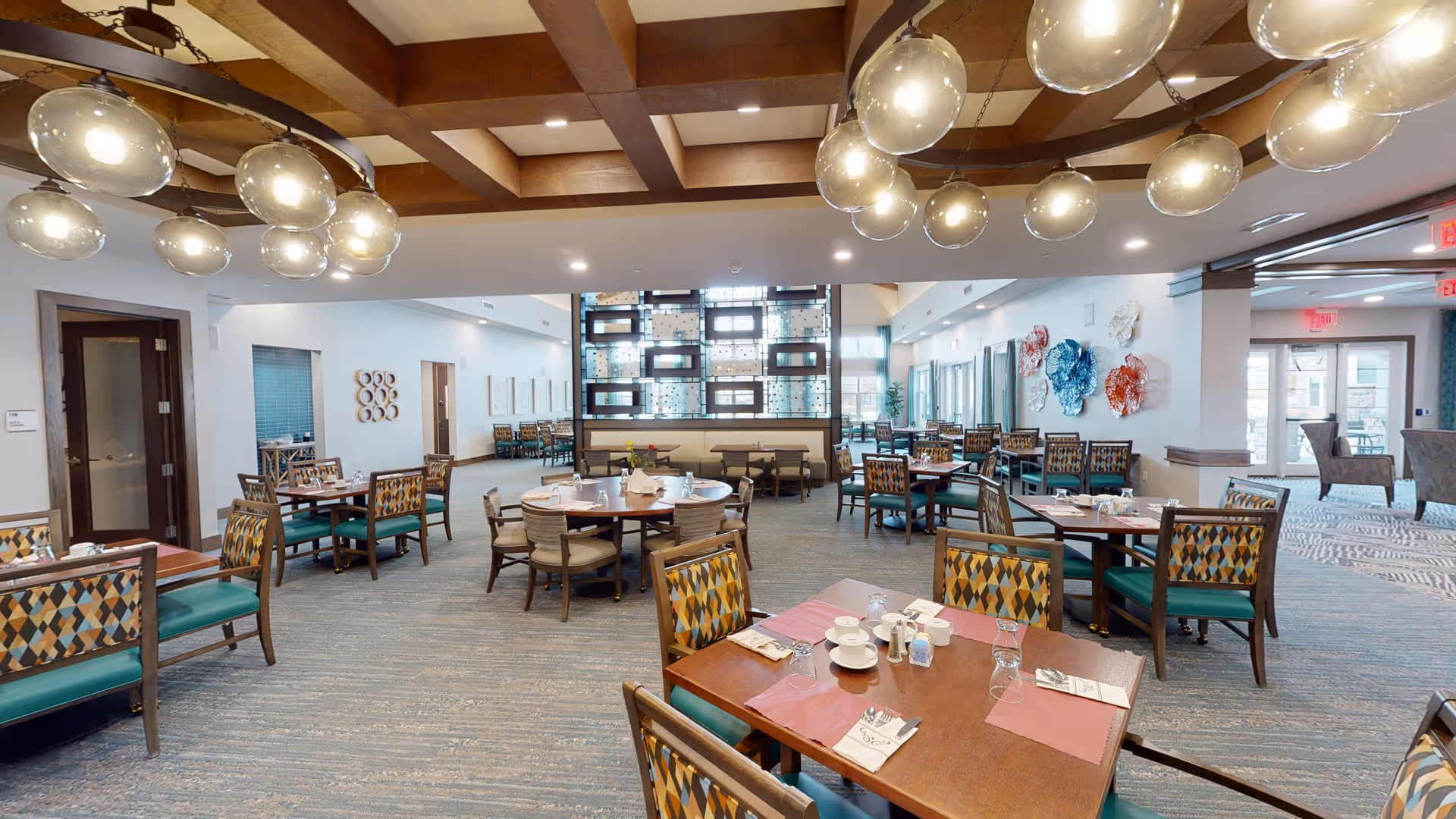 A spacious dining room in a senior living facility with multiple wooden tables and chairs arranged neatly. The chairs have patterned upholstery with green cushions. The room features a coffered ceiling with modern circular light fixtures. Decorative wall art and a partition with glass blocks are visible in the background. The room is well-lit with natural light coming through large windows and doors.