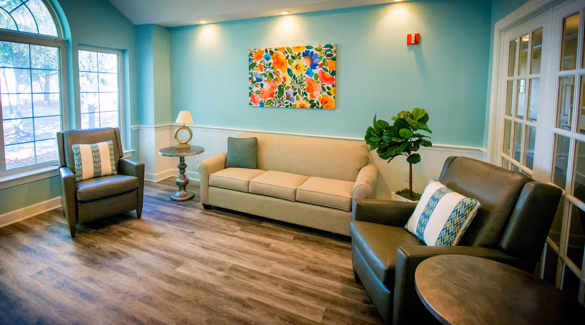 Sunlit seating lounge with a beige sofa, two leather armchairs, side tables, a potted plant, and colorful floral artwork on a teal wall.