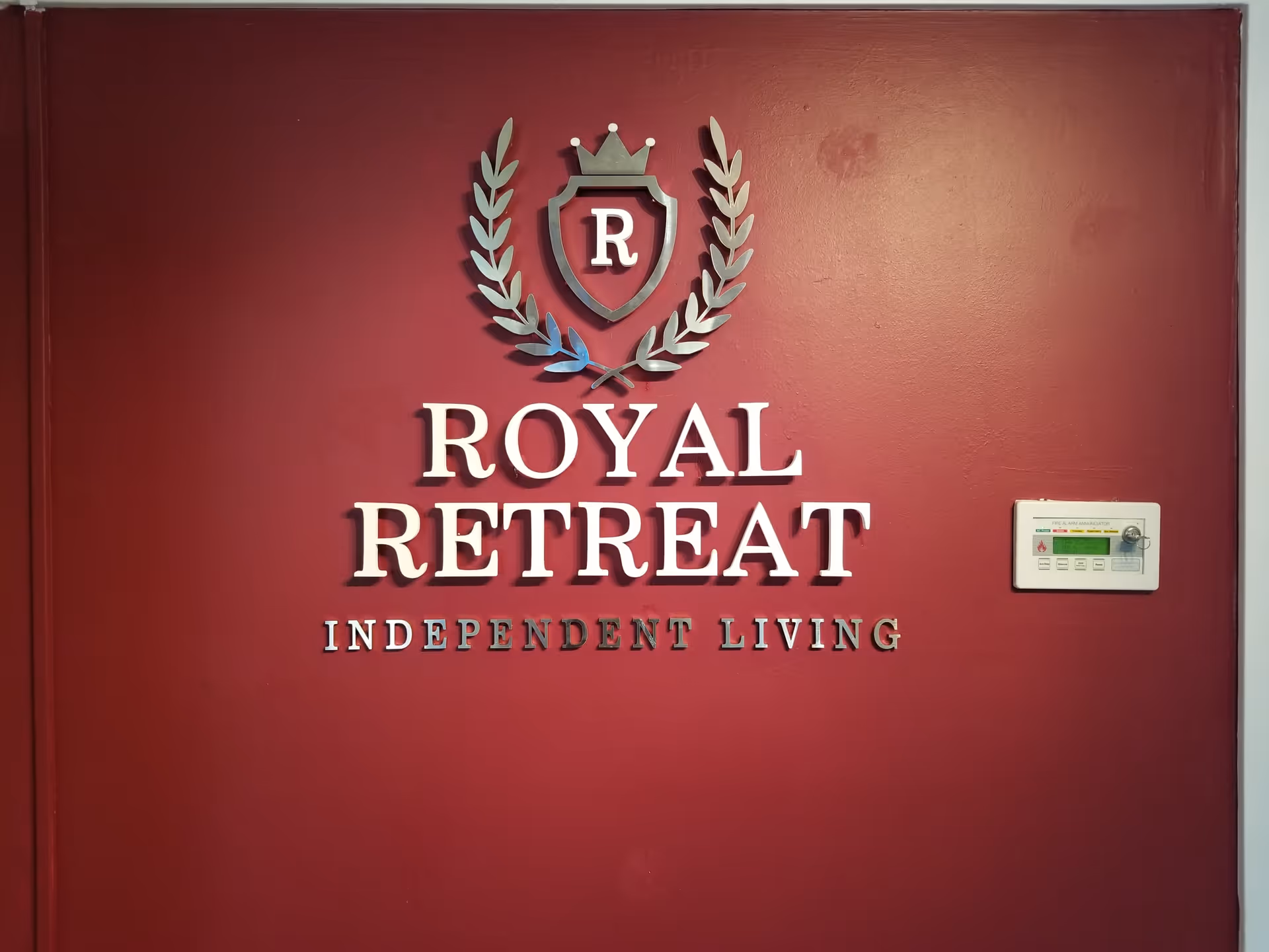 A maroon wall with a silver emblem featuring a crown and laurel leaves above the text 'ROYAL RETREAT INDEPENDENT LIVING'. To the right of the text is a fire alarm annunciator panel.