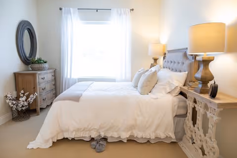 A bright and cozy bedroom with a neatly made bed featuring white bedding and multiple pillows. There are two wooden nightstands on either side of the bed, each with a lamp. A wooden dresser with a round mirror above it is positioned against the wall to the left. White curtains frame a window letting in natural light. A pair of slippers is placed on the floor near the foot of the bed.