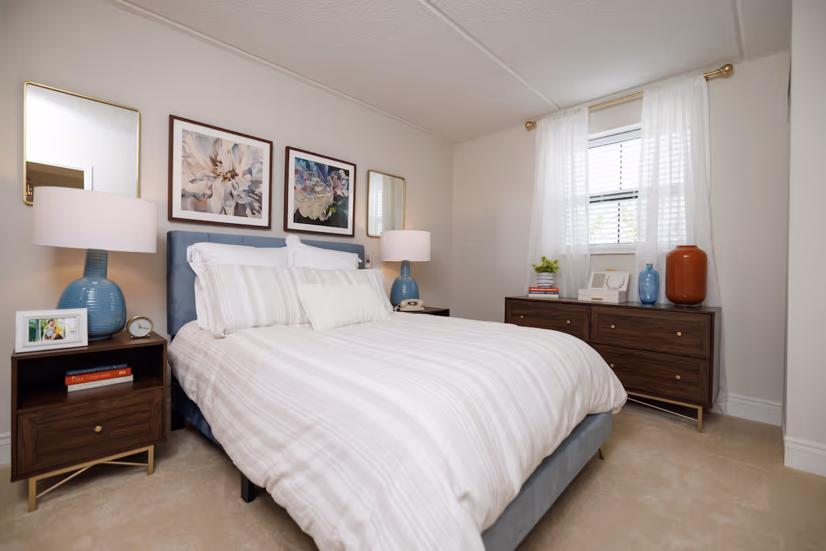 A neatly made bed with white and light gray striped bedding in a bedroom. The bed has a blue upholstered headboard and is flanked by two wooden nightstands, each with a blue lamp and decorative items. Above the bed are two framed floral artworks and two mirrors. A wooden dresser with decorative vases, books, and a framed photo is positioned under a window with sheer white curtains.