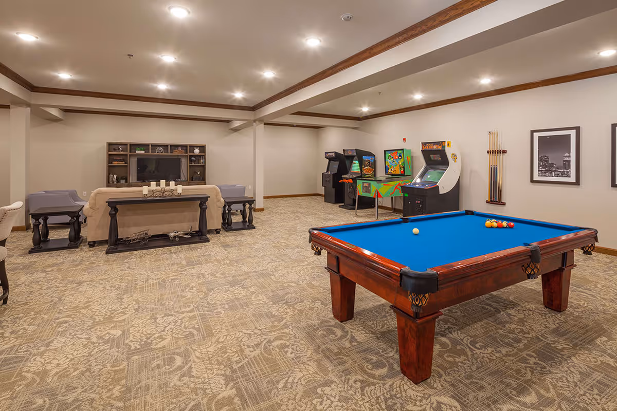 A spacious game and lounge room featuring a blue felt pool table in the foreground, several arcade game machines along the back wall, and a seating area with a beige sofa and two chairs facing a TV on a wooden entertainment center. The room has beige walls, patterned carpet, recessed lighting, and framed black and white photos on the wall.