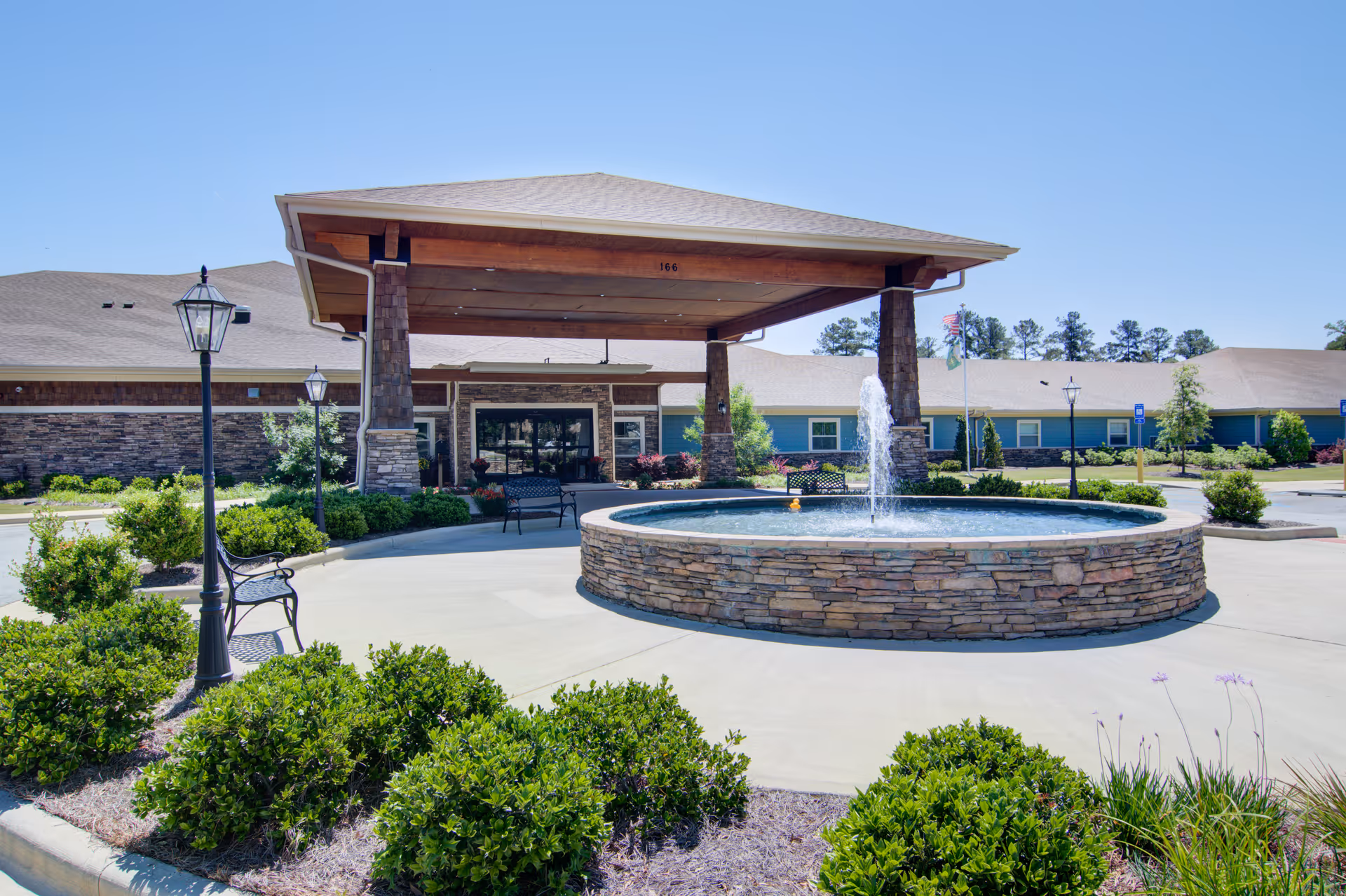 Exterior view of Oaks at Oakland Plantation featuring a covered entrance with stone pillars, a circular stone fountain with water spraying upwards, benches, lamp posts, and landscaped greenery under a clear blue sky.