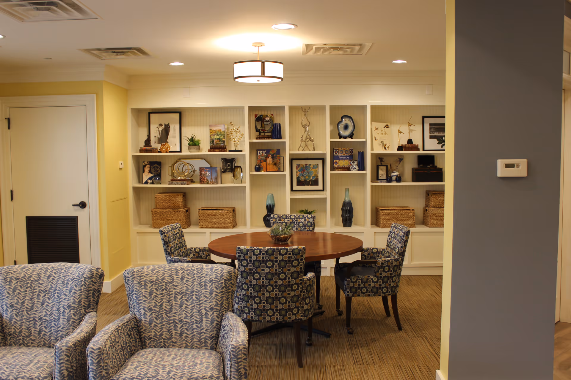A cozy interior room with a round wooden table surrounded by four patterned chairs. Behind the table is a built-in white shelving unit filled with decorative items, books, baskets, and framed pictures. Two patterned armchairs are partially visible in the foreground. The walls are painted yellow and gray, and a ceiling light fixture is centered above the table.