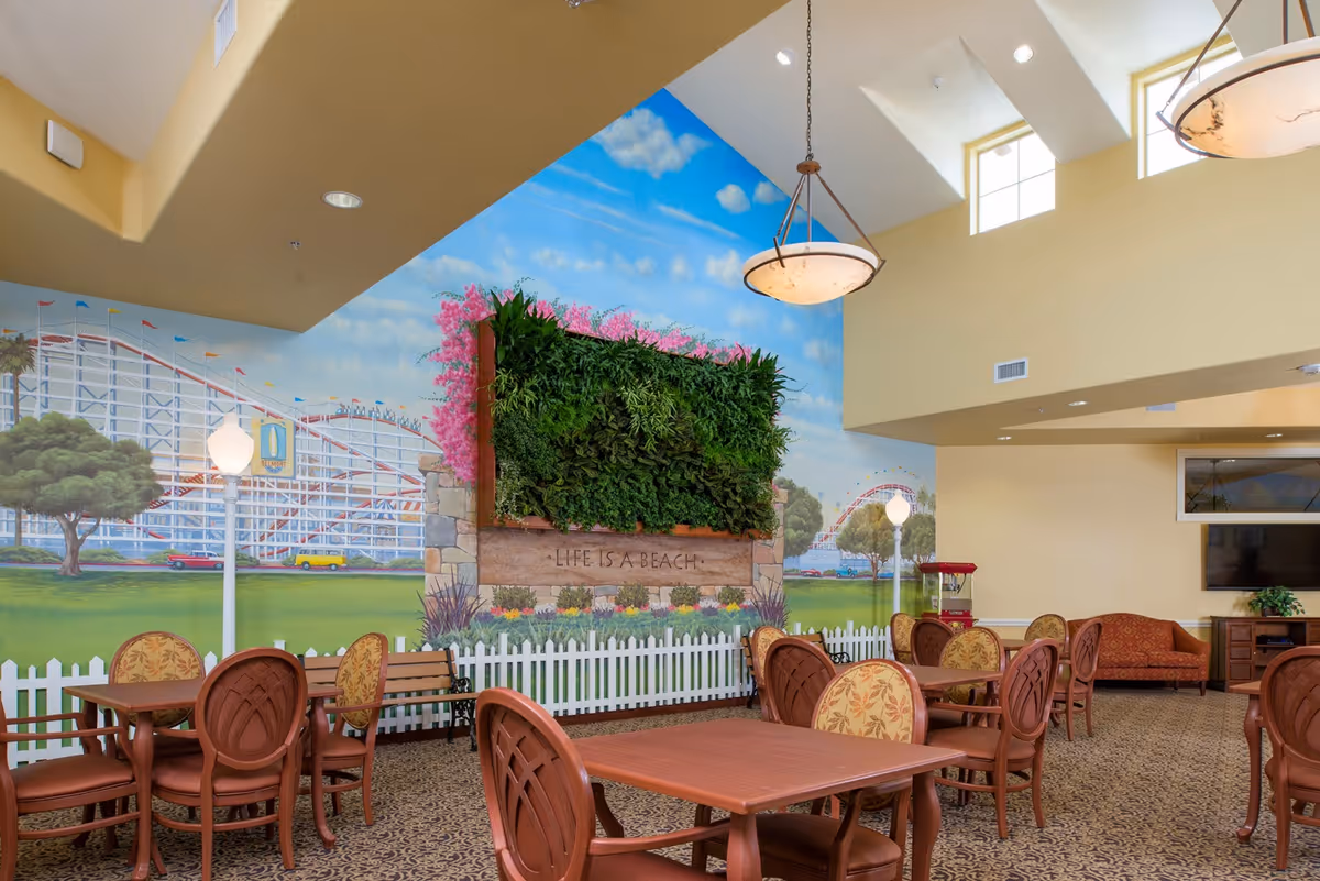 A bright and spacious common area with multiple wooden tables and chairs arranged on a patterned carpet. The walls feature a large mural depicting a roller coaster, trees, and a blue sky with clouds. A vertical garden with green plants and pink flowers is mounted on the wall above a white picket fence, with the phrase 'LIFE IS A BEACH' written on a wooden panel beneath it. The room has high ceilings with hanging light fixtures and windows near the ceiling allowing natural light in. There is a red popcorn machine and a TV mounted on the wall in the background.