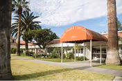 Exterior view of Villa Health Care Center showing the entrance with an orange awning displaying the facility's name and the year 1963. The building is surrounded by palm trees and a well-maintained lawn under a partly cloudy sky.
