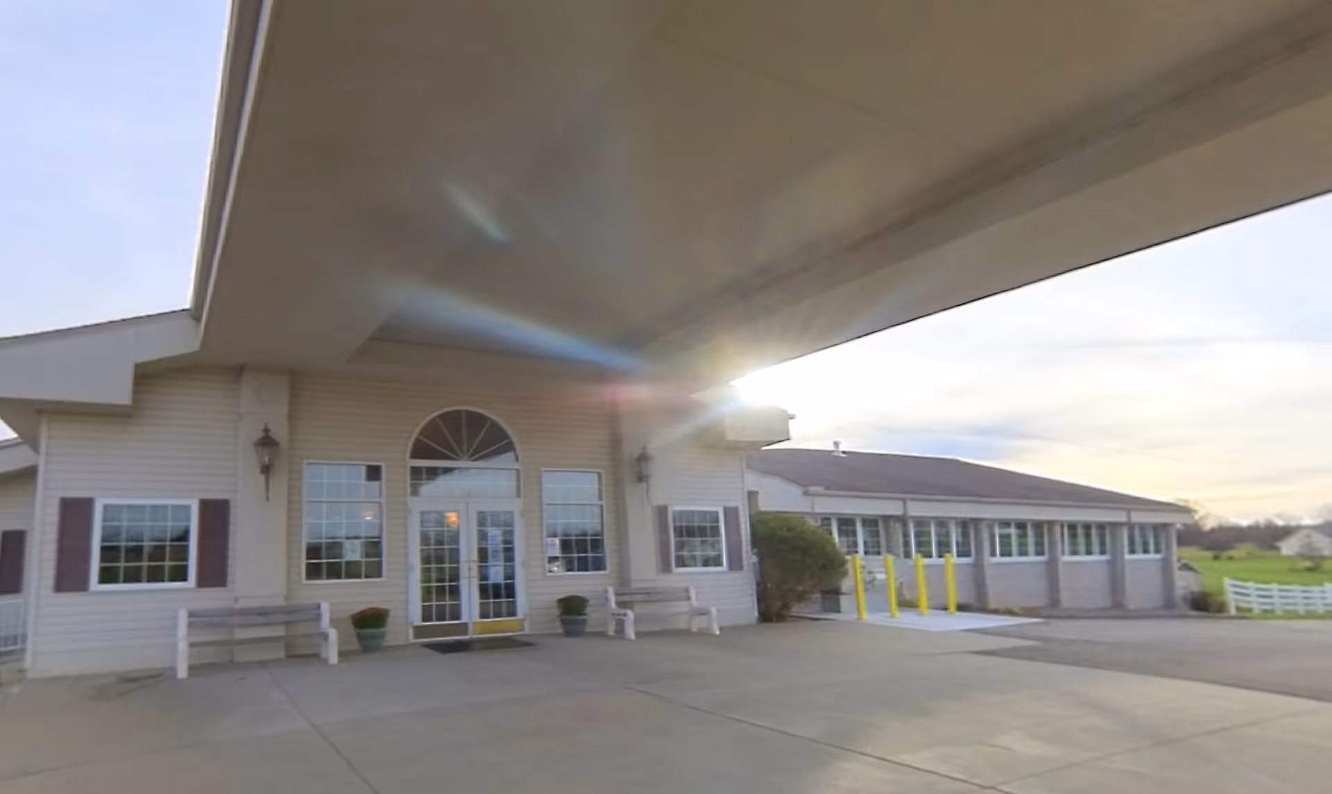 Exterior view of the entrance to a senior living facility with a covered drop-off area, large windows, benches, and potted plants near the doorway. The building is light-colored with a sloped roof and surrounded by open space and a fence in the background.