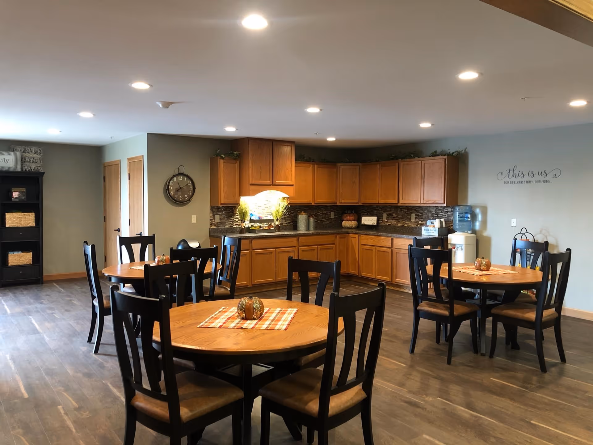 A dining area with round wooden tables and black chairs, each table decorated with a small centerpiece on a plaid placemat. In the background, there is a kitchen area with wooden cabinets, a countertop, a water dispenser, and a wall clock. The walls are painted light gray and there is a decorative wall decal that reads 'this is us, our life, our story, our home.'