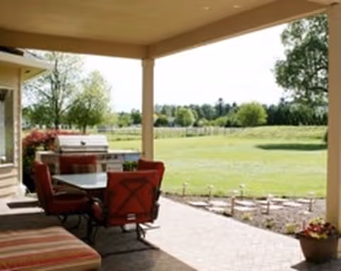 Covered patio with a dining table, red chairs and a grill overlooking a grassy yard and trees.