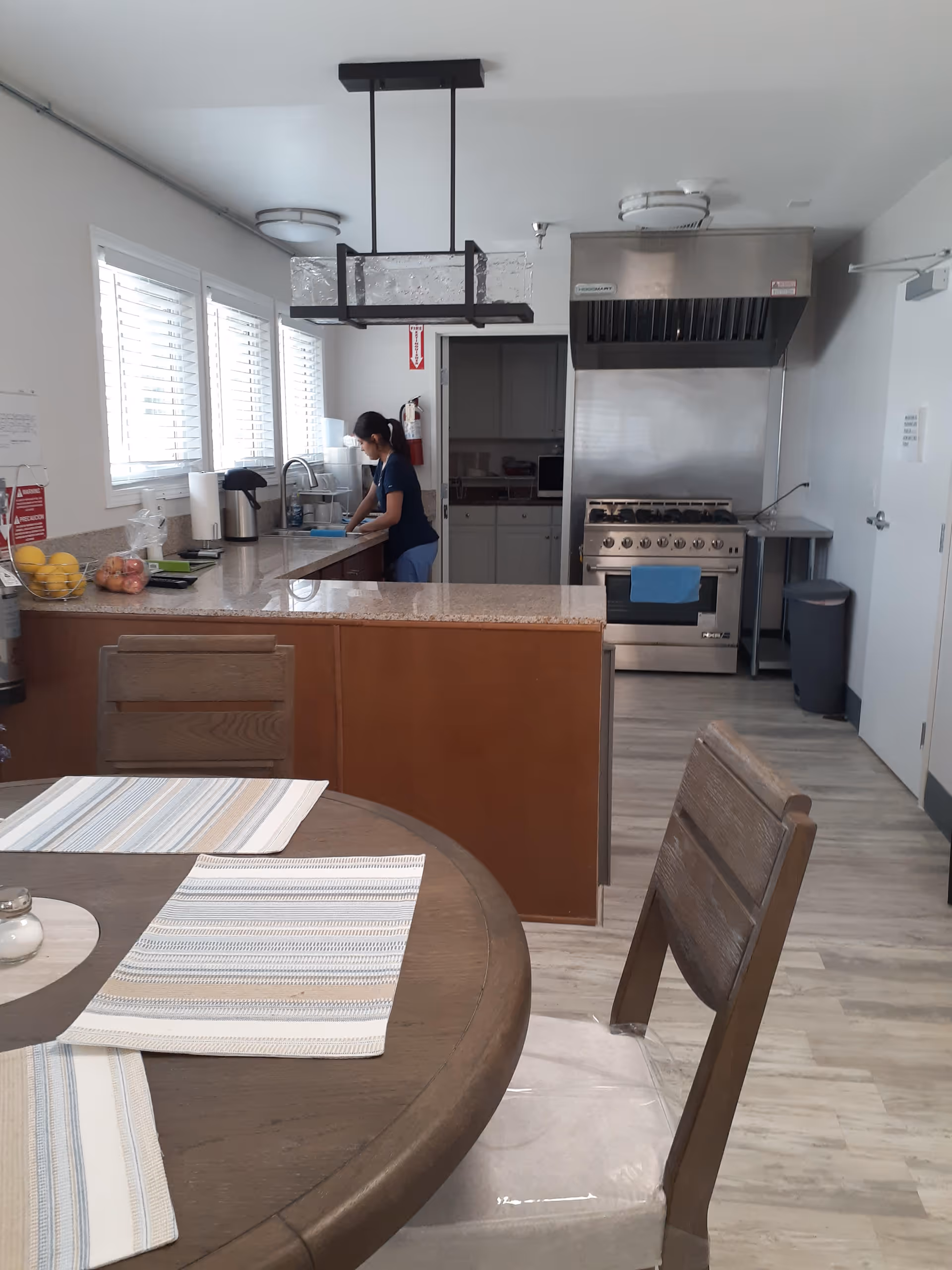A kitchen area with a woman washing dishes at the sink. The kitchen has a large stainless steel stove with an exhaust hood, a countertop with fruit baskets, and a dining table with placemats and wooden chairs in the foreground. The room is well-lit with natural light coming through the windows with white blinds.