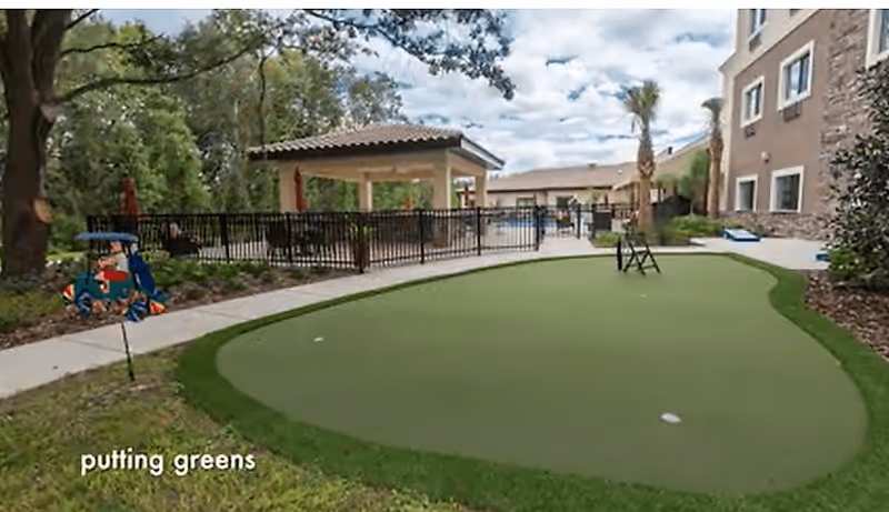 Outdoor putting green area with a small gazebo and pool fenced in the background, surrounded by trees and a multi-story building.