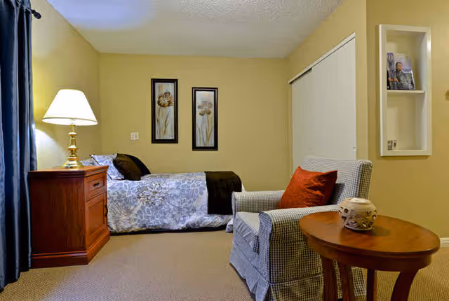 A cozy bedroom with a single bed, wooden nightstand and lamp, patterned armchair with a red pillow, and framed floral artwork on the wall.
