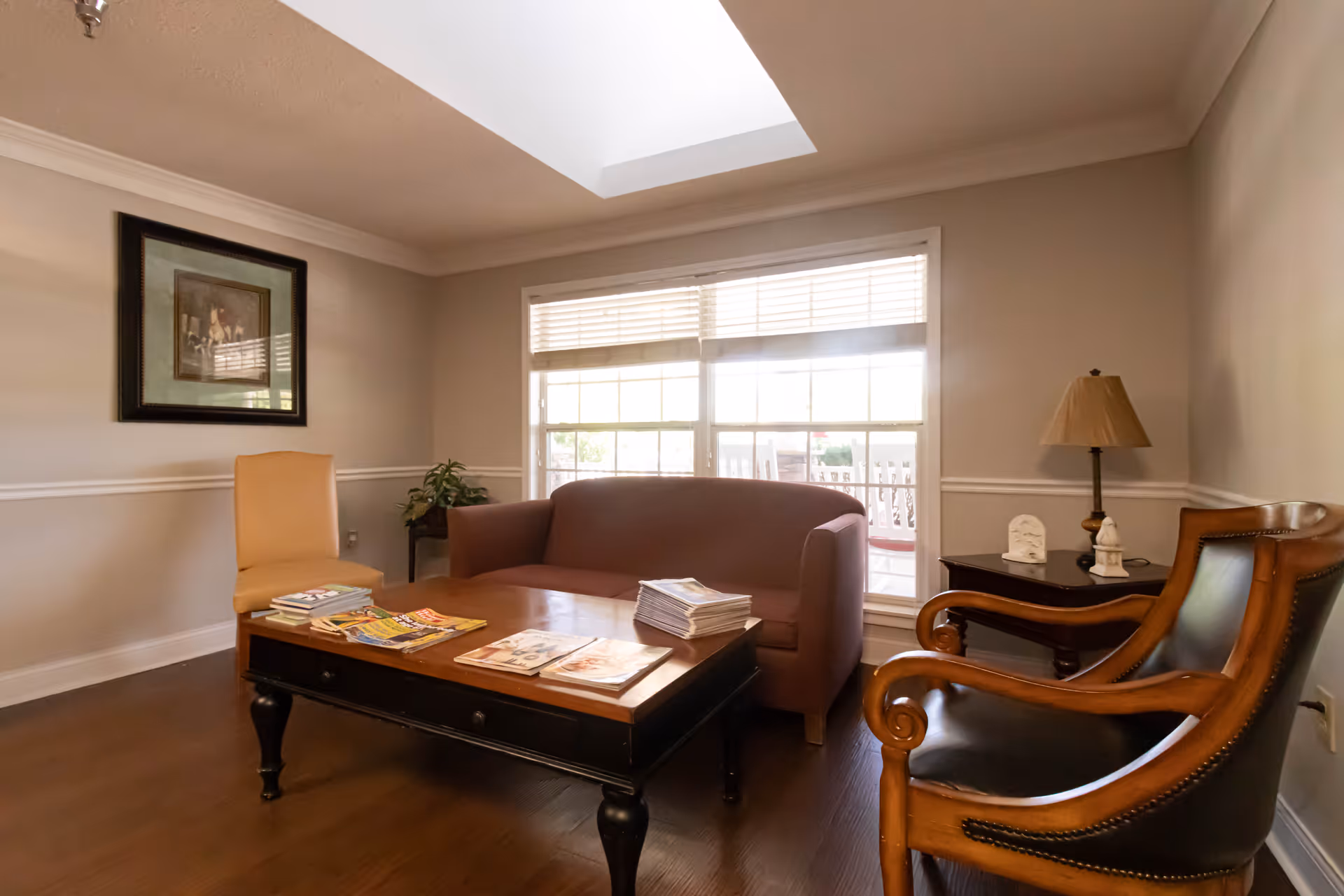 A cozy living room with a brown sofa, a wooden coffee table with magazines on it, a beige chair, and a wooden armchair with black leather upholstery. There is a side table with a lamp and decorative items, a framed picture on the wall, and a large window with blinds letting in natural light.