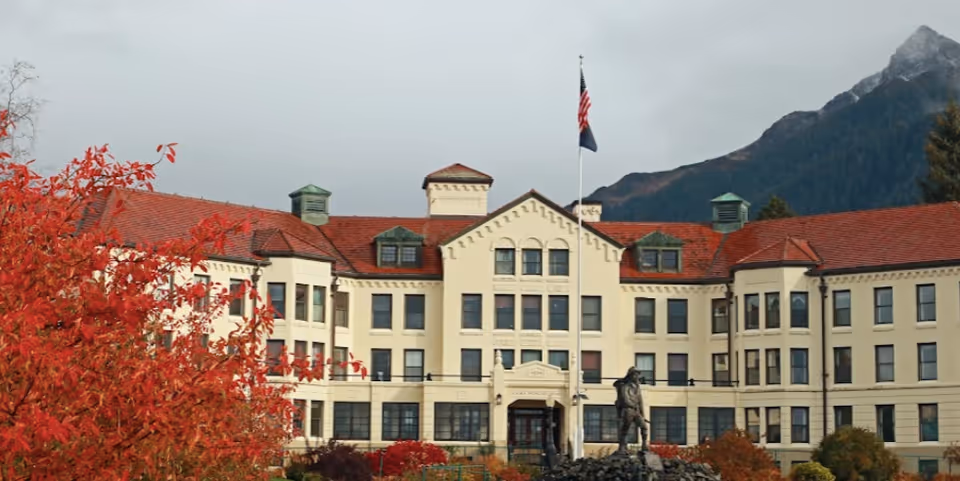 Front exterior view of the Pioneers' Home Sitka building with a statue in front, an American flag on a flagpole, red autumn foliage on the left, and mountains in the background under a cloudy sky.