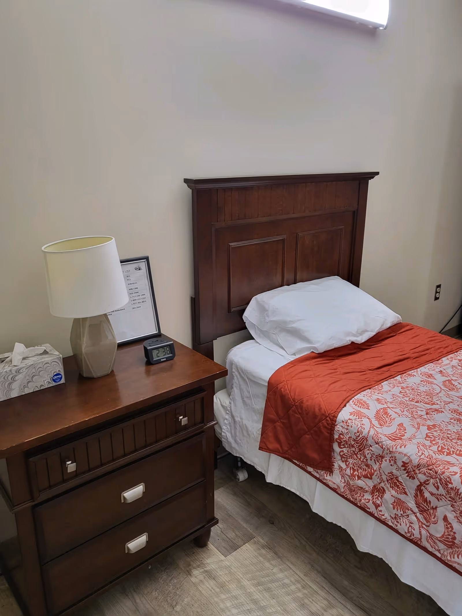 A neatly made single bed with a wooden headboard, white pillow and sheets, and a red patterned blanket. Next to the bed is a wooden nightstand with a beige lamp, a small digital clock, a box of tissues, and a framed document. The room has light-colored walls and wood flooring.