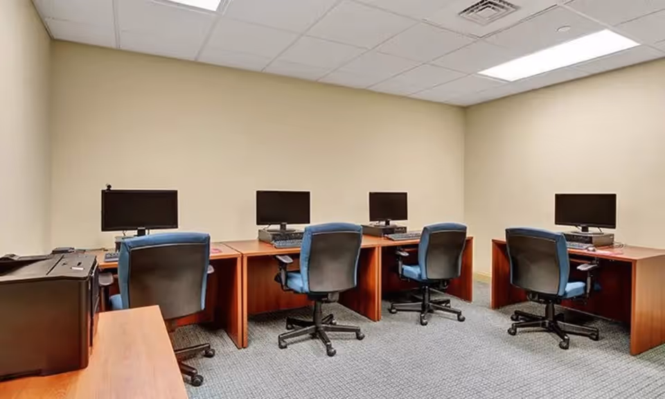Small office computer room with four wooden desks, monitors and blue swivel chairs under a drop ceiling.