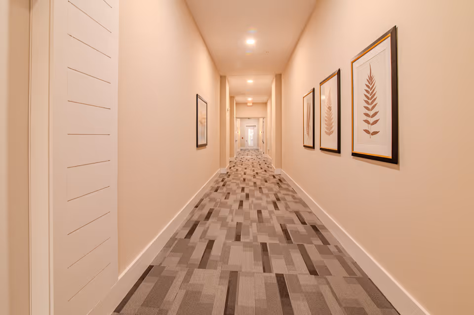 A long, well-lit hallway with beige walls and a patterned carpet floor. The hallway features framed botanical prints on the right wall and a door at the far end with an exit sign above it.