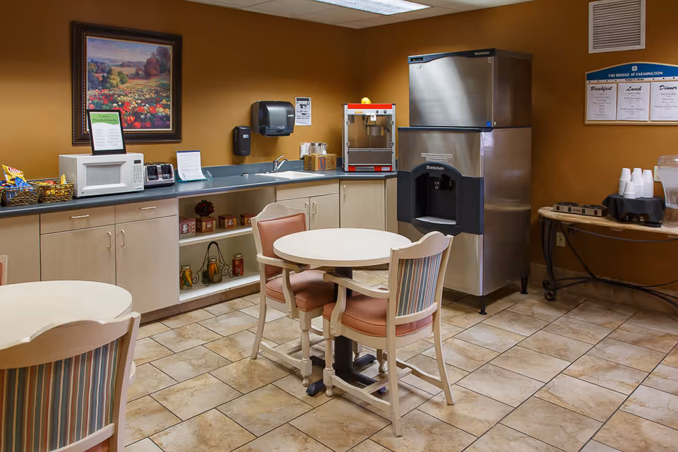 Small dining area with two round tables and chairs, a countertop with a microwave, popcorn machine, and an ice dispenser. The walls are painted brown and there is a framed landscape painting and a menu board on the wall.