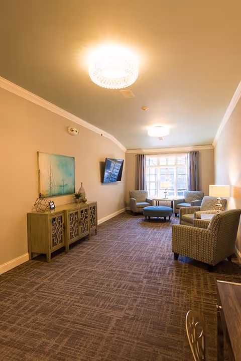 A well-lit living room area with beige walls and carpeted floor. The room features a decorative sideboard with artwork and small plants, a wall-mounted flat-screen TV, and a seating area with four patterned armchairs arranged around a round ottoman near a large window with curtains. Two ceiling lights illuminate the space.