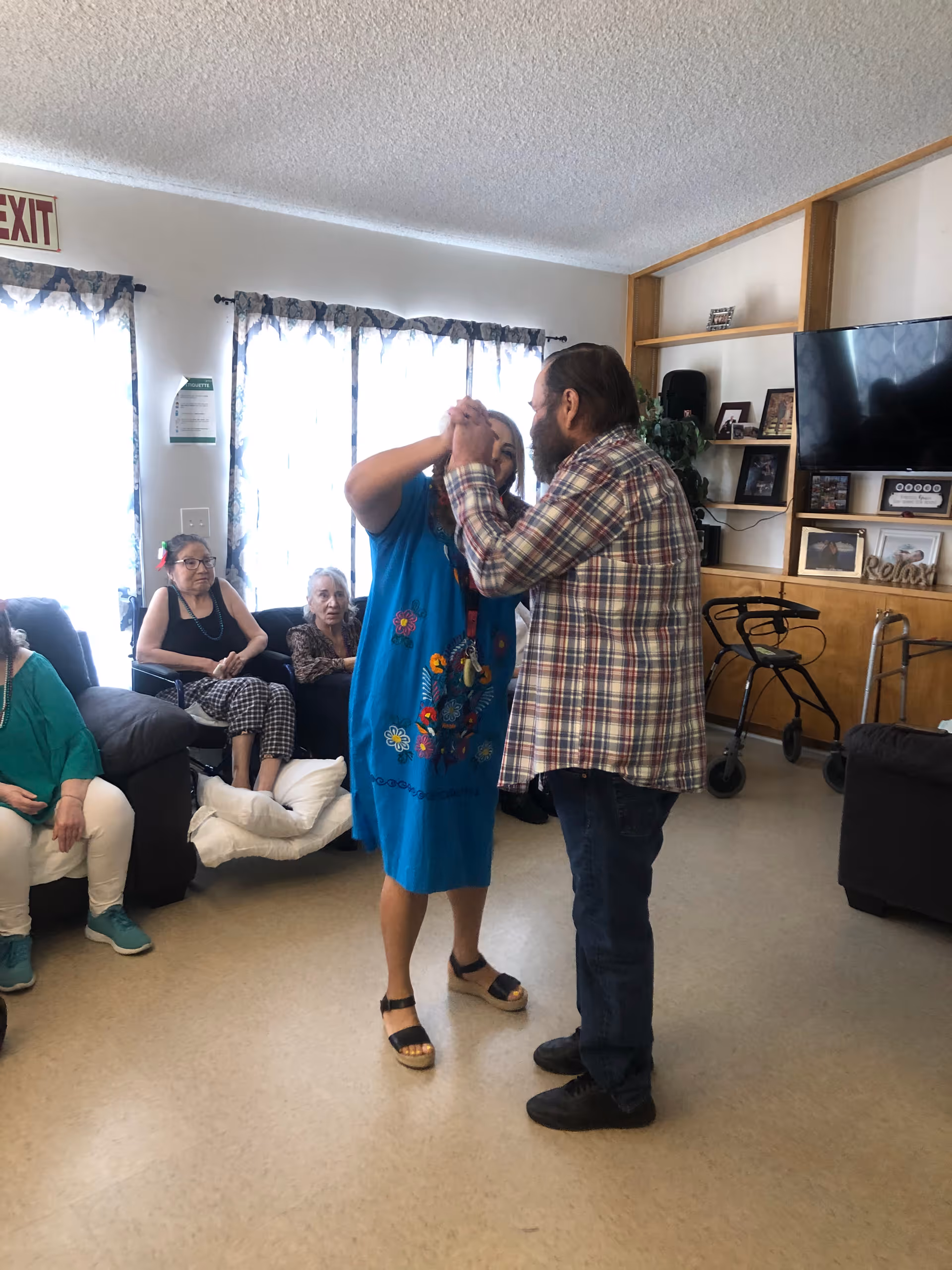 Two people dancing together in a living room area of an assisted living facility. Several elderly individuals are seated on couches and chairs in the background, watching the dancers. The room has a television, shelves with framed photos, and a walker near the wall.