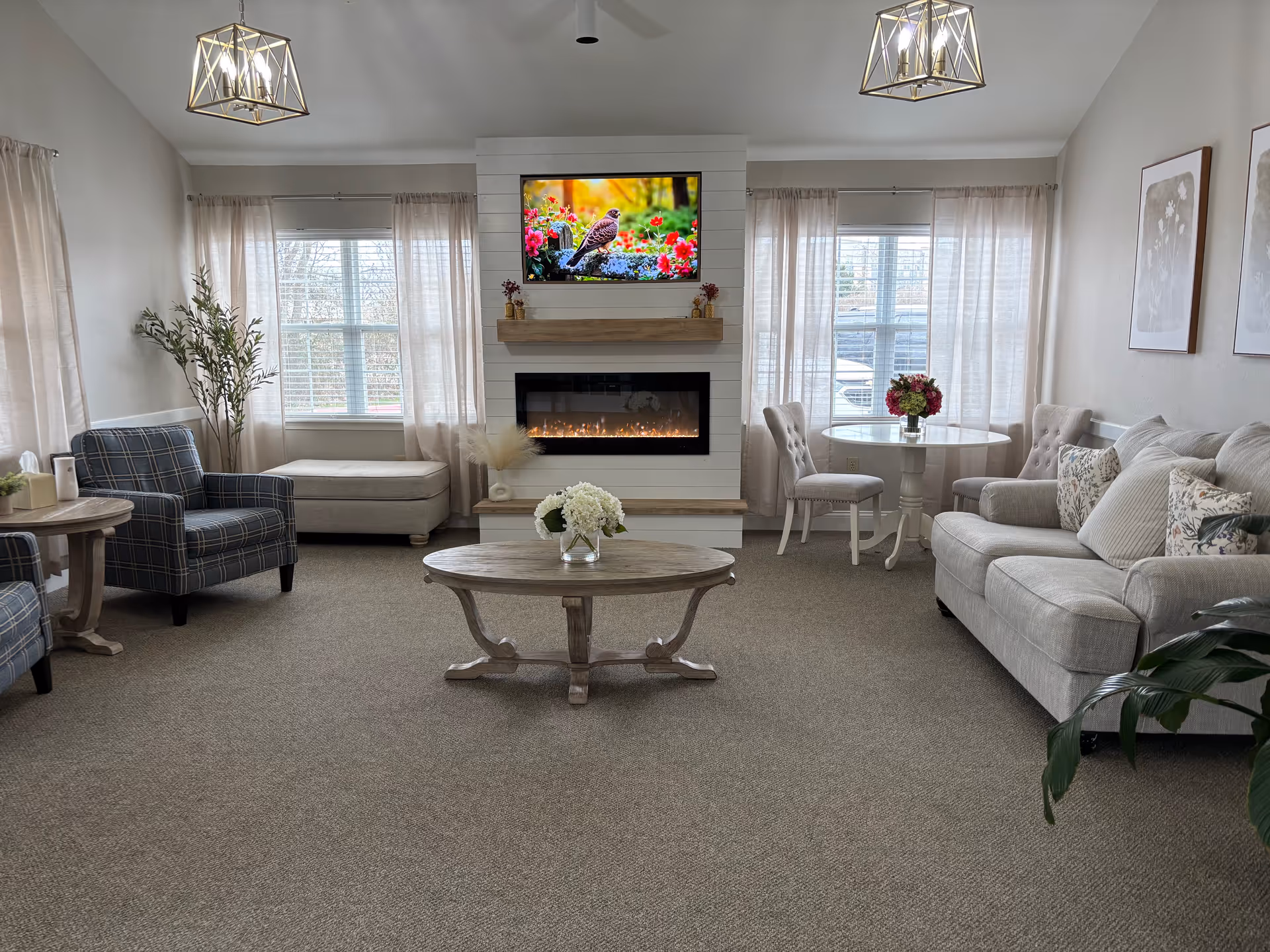 A cozy living room with a beige carpet, a light gray sofa with patterned pillows, two plaid armchairs, a round wooden coffee table with a flower vase, and a small round dining table with two chairs. The room features a modern electric fireplace with a TV mounted above it displaying a bird and flowers. Large windows with sheer curtains allow natural light to fill the space, and two geometric pendant lights hang from the ceiling.