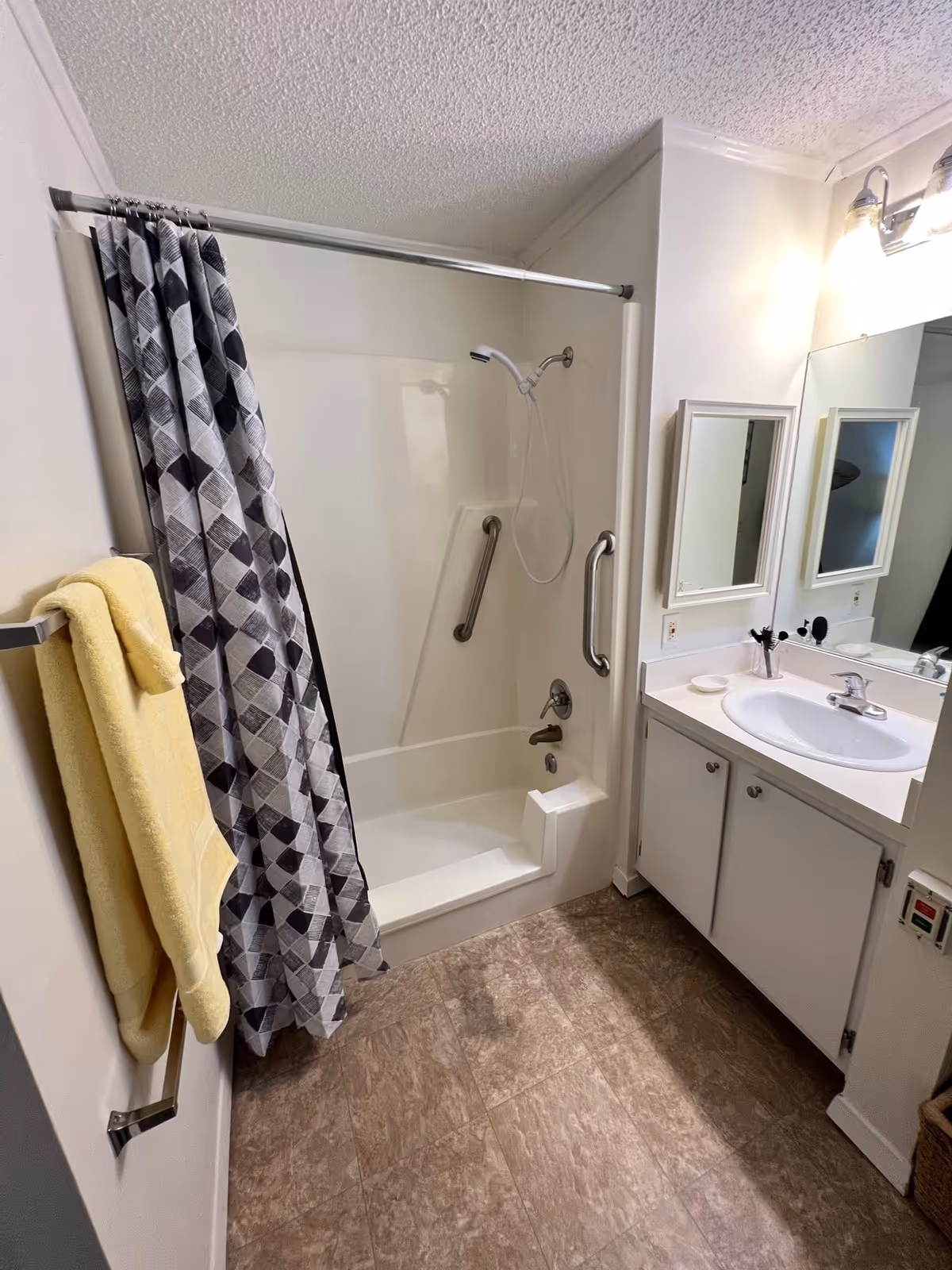 A clean bathroom with a white bathtub and shower combination featuring a handheld showerhead and two grab bars. A gray and white patterned shower curtain is partially drawn. A yellow towel hangs on a towel rack on the left wall. To the right is a white vanity with a sink, a mirror, and a light fixture above. The floor has a brown tile pattern.