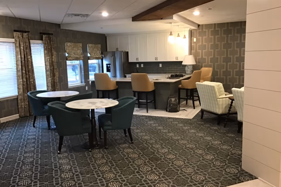 Interior view of a senior living facility common area featuring a kitchen with white cabinets, a refrigerator, and a countertop with four tan bar stools. In the foreground, there are two round tables each surrounded by teal upholstered chairs. To the right, there are two cream-colored armchairs with a patterned wall behind them. The room has patterned carpet and windows with curtains and blinds.