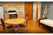 Small nursing-home room with a kitchenette, round dining table and chairs, and a single bed against the wall.