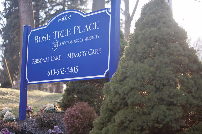 Blue outdoor sign for Rose Tree Place reading "Personal Care | Memory Care" standing in a landscaped yard.