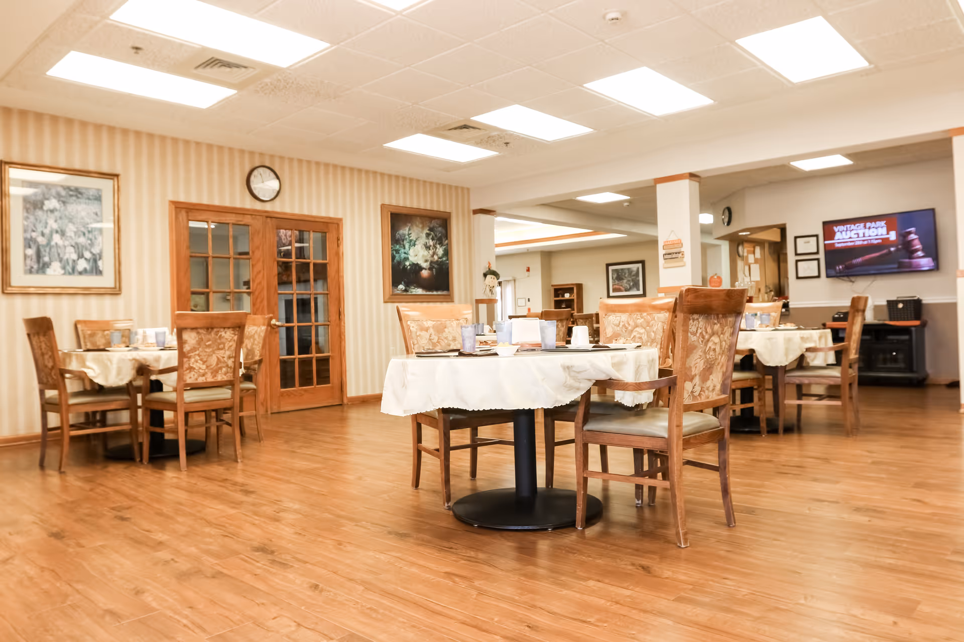 A dining room in a senior living facility with several round tables covered with white tablecloths and set with cups and plates. The room has wooden chairs with floral upholstery, wood flooring, and beige striped wallpaper. There are framed paintings on the walls, a clock above wooden double doors, and a television screen displaying 'Vintage Park Auction' in the background.