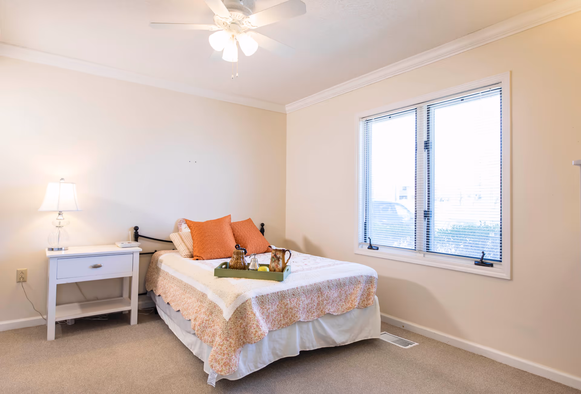 A simple bedroom with a bed covered in a floral quilt and two orange pillows. A white nightstand with a lamp and a phone sits next to the bed. There is a window with blinds on the right wall and a ceiling fan with a light above.