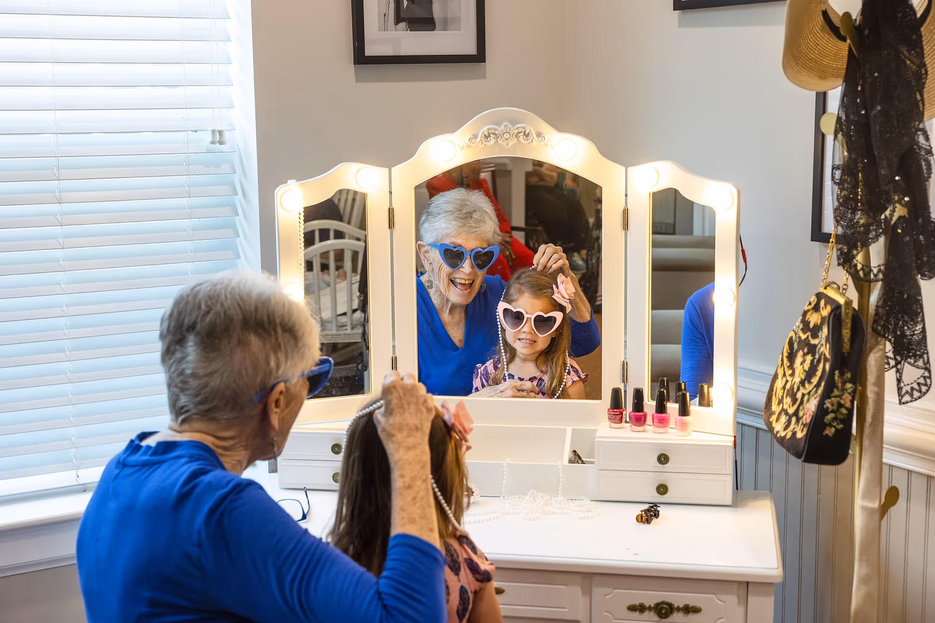 An elderly woman and a young girl wearing heart-shaped sunglasses are sitting at a white vanity with a tri-fold mirror surrounded by lights. The elderly woman is smiling and adjusting the girl's hair while the girl looks at their reflection. On the vanity are several bottles of nail polish and a string of pearls. A window with blinds is on the left, and a coat rack with hats and bags is on the right.