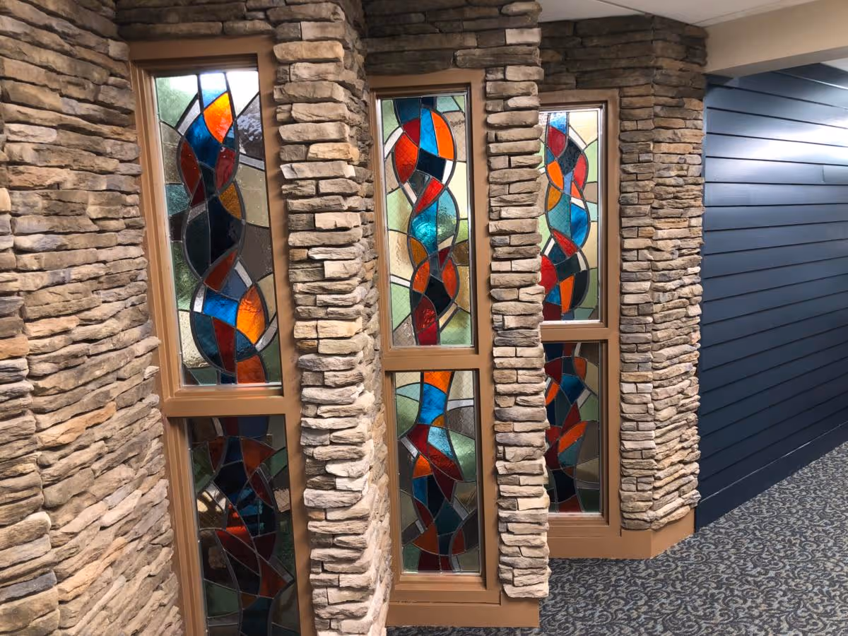 Interior view of a hallway featuring decorative stained glass windows framed by stone columns on the left side and a blue paneled wall on the right side, with patterned carpet flooring.