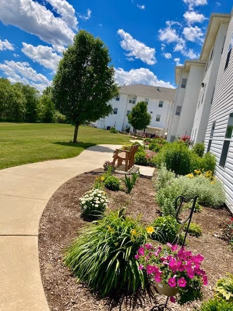 A sunny outdoor garden area at Supportive Living of Washington featuring a curved concrete walkway, green grass, a variety of colorful flowers and plants, a tree, and wooden benches along the path. The white exterior of the building is visible on the right side under a blue sky with scattered clouds.