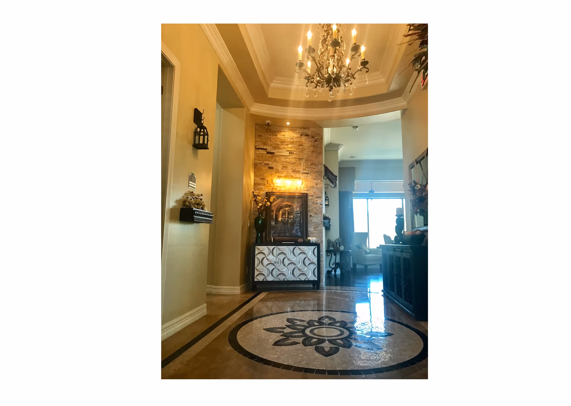 An elegant interior hallway with a decorative circular mosaic tile floor, a chandelier hanging from a tray ceiling, and a console table against a stone accent wall. The hallway leads into a bright living area with large windows and comfortable seating.