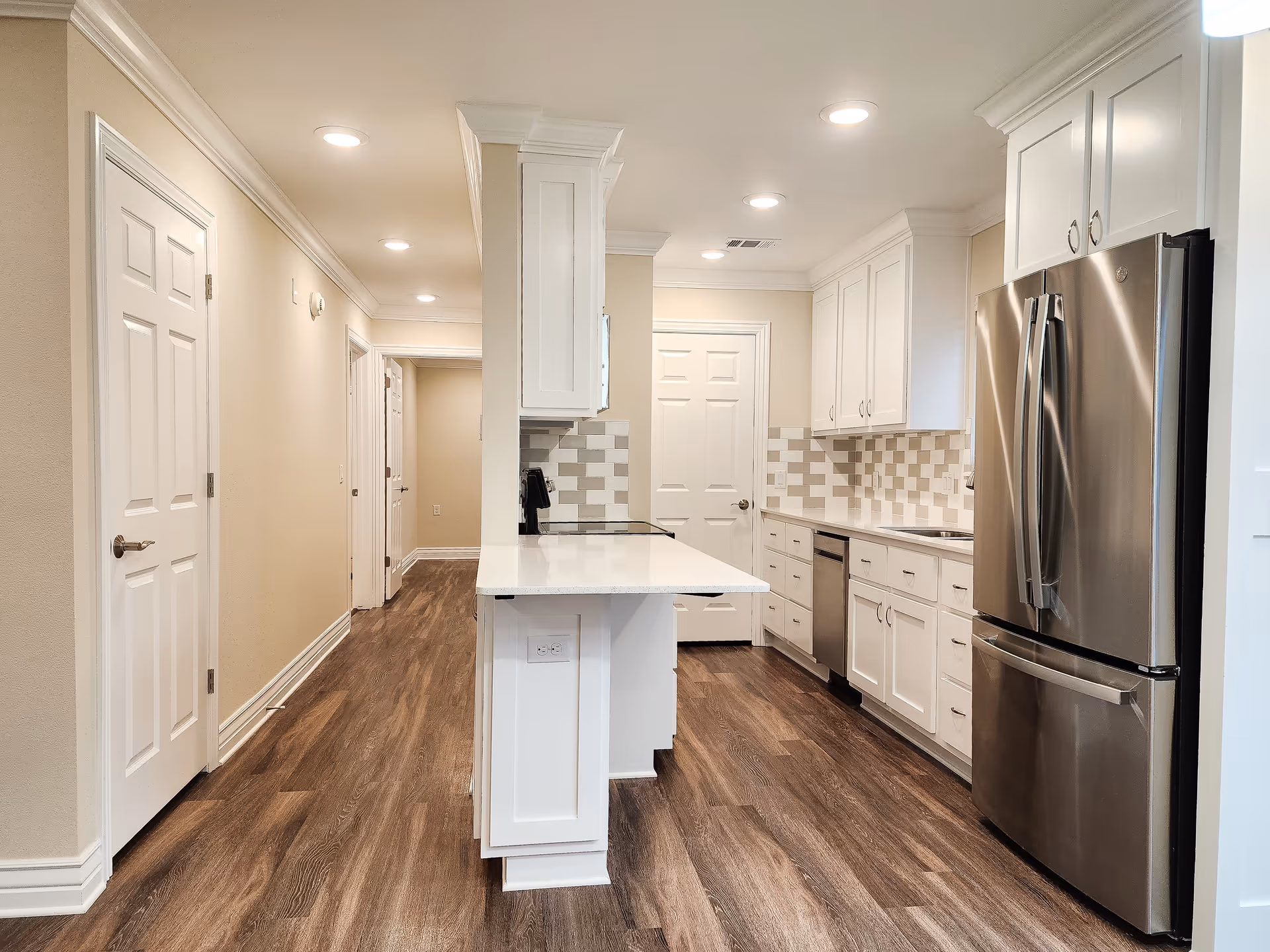 A modern kitchen with white cabinets, a stainless steel refrigerator, dishwasher, and a white countertop island. The floor is wood with a dark finish, and the walls are painted beige. Recessed ceiling lights illuminate the space, and there is a hallway visible to the left.